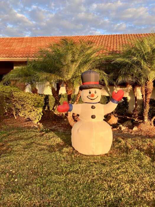 Uppblåsbar snögubbe med hatt och röda handskar står på en gräsmatta framför en byggnad med terrakottatak och omgiven av palmer.