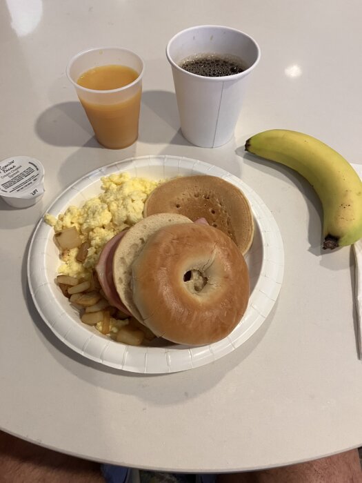 Frukost med bagel, äggröra, potatis, pannkaka, apelsinjuice, kaffe, grädde och banan på ett vitt bord.