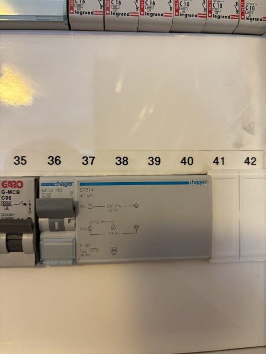 Transformator installerad i ett elskåp, märkt Hager ST314, omgiven av strömbrytare. Nummermarkeringar synliga nedanför installationen.