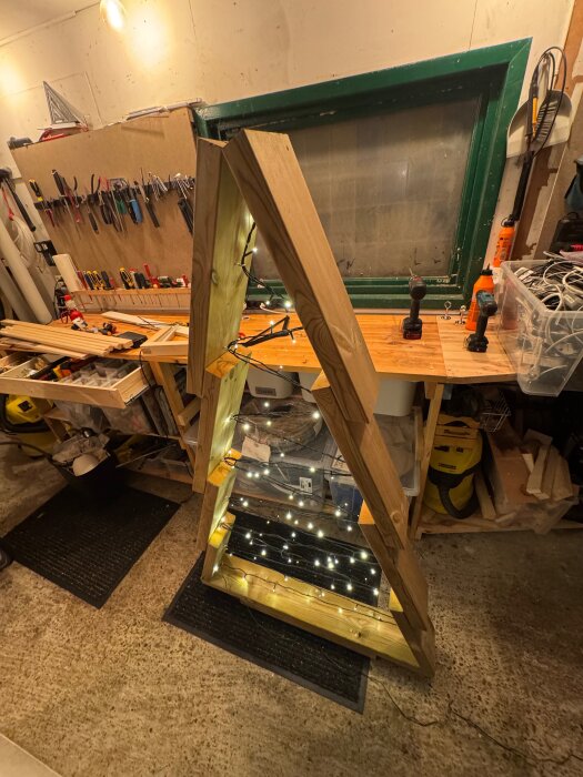 Trekantig trästruktur dekorerad med ljusslingor i en verkstad med verktyg och arbetsbänk i bakgrunden.