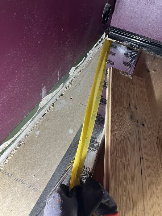 Mätningsband påträffar uppreglat golv med träplintar, träreglar och fuktspärr under parkett i källare, mot en lila vägg.
