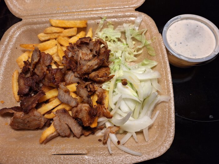 Gyrostallrik med pommes frites, gyroskött, sallad och lök i en kartonglåda, med en plastburk vit sås vid sidan av.