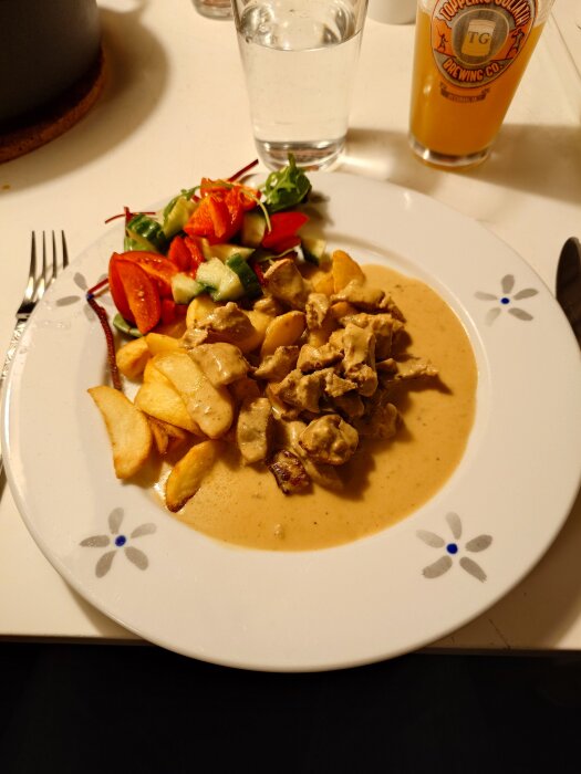 Fläskfilégryta med klyftpotatis, serverad med sallad innehållande tomater och gurka, på en vit tallrik. Glas med dryck i bakgrunden.