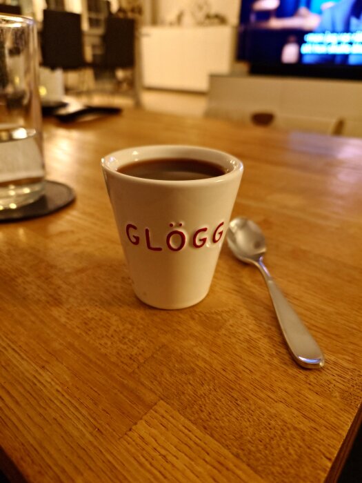 En vit mugg med "GLÖGG" tryckt i rött, fylld med glögg, står på ett träbord bredvid en tesked.