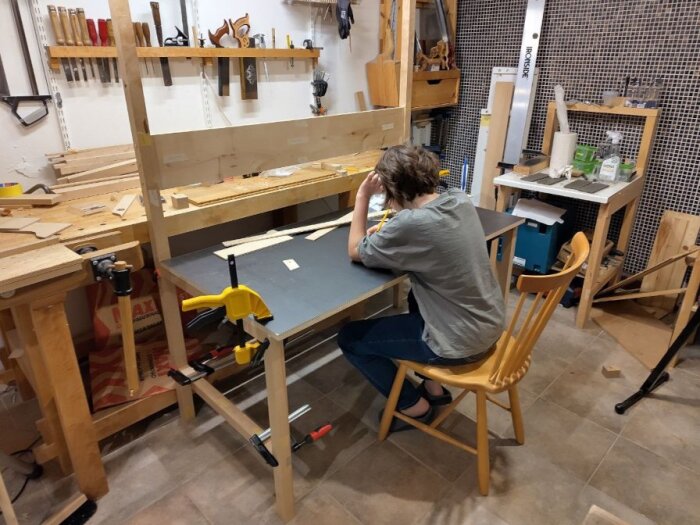 Person sitter vid arbetsbord i verkstad, mäter höjden på en bänkskiva och använder papper med anteckningar. Verktyg och trädelar syns på borden.