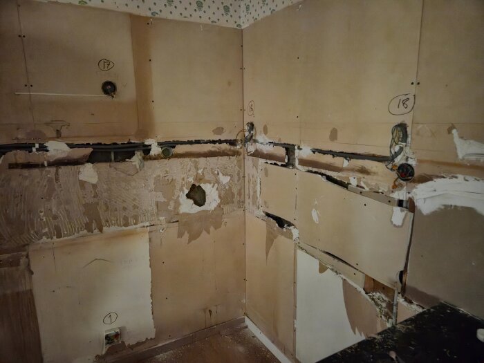 Rivet kök med trasiga väggar och exponerade ledningar, tecken på slitna och oanvändbara material efter renovering, delvis avskalat tapet.