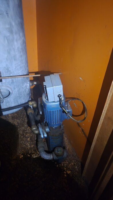 Blå vattenpump inuti ett hus med orange vägg i bakgrunden, visar sladdar och anslutna rör.