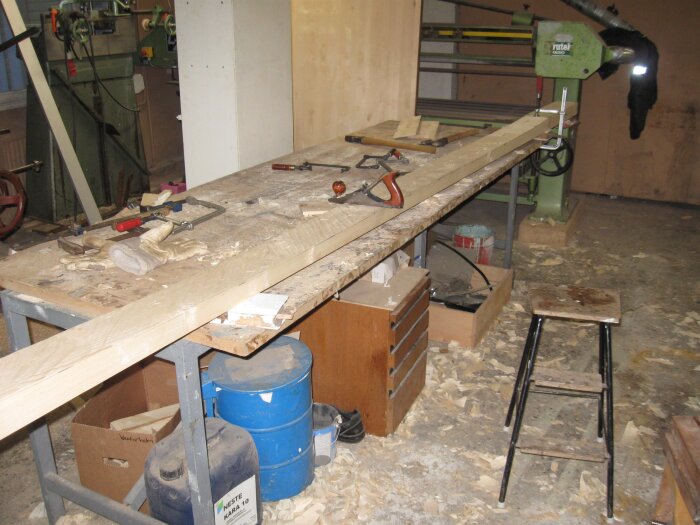 Arbetsbänk i verkstad med sågspån; verktyg, en träbit under bearbetning och en maskin i bakgrunden.