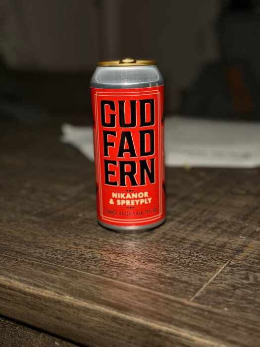 En röd burk med texten "Gudfadern," en starköl från Nikanor & Spreply, står på ett träbord.