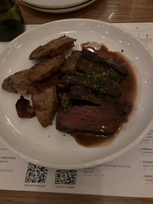 Tallrik med stekt kött och stekta potatisklyftor, täckt med sås och gräslök. I bakgrunden syns en del av en meny och en flaska.