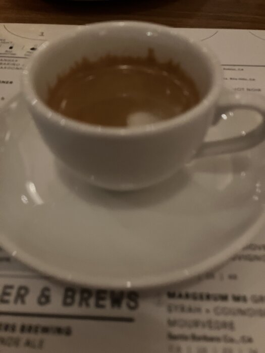 En vit kopp med kaffe på ett bord, med en suddig meny i bakgrunden.
