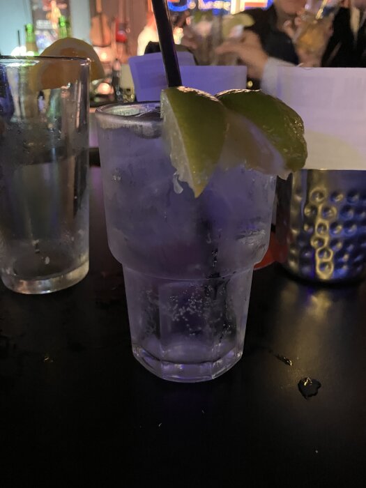 Ett glas med is och limeklyftor samt sugrör på ett bord i en bar, med fler drycker och människor i bakgrunden.