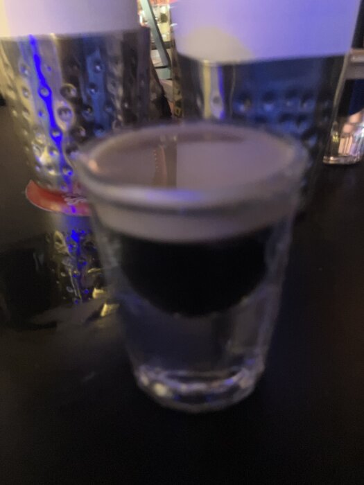 Ett snapsglas med mörk vätska på ett bord, omgiven av metallmuggar i bakgrunden. Dugligt oskarp bild med blåaktig belysning.