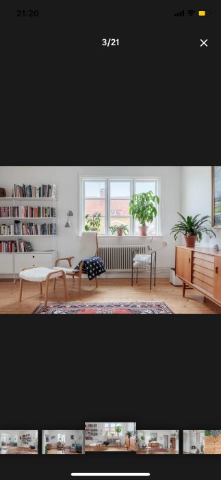 Ljust vardagsrum med fåtölj, bokhylla och växter på fönsterbräda, sett genom ett stort fönster.