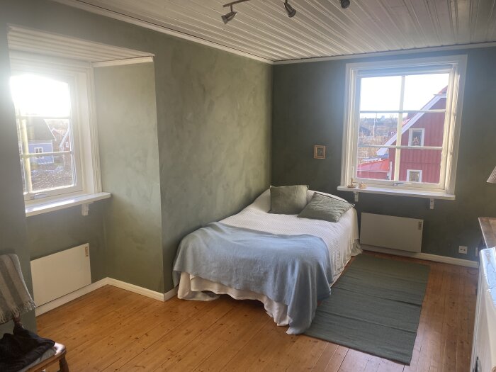 Sovrum med fönster och träpanelstak, grönmålade väggar och trägolv. En bäddad säng står vid fönstret med grå filt och kuddar.