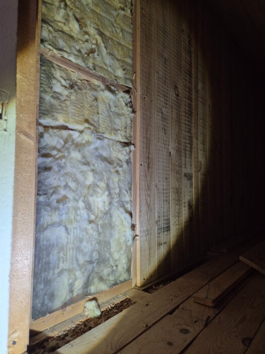 Exposed wooden wall structure with visible insulation, showing a gap between panels and a wooden beam in an attic under renovation.