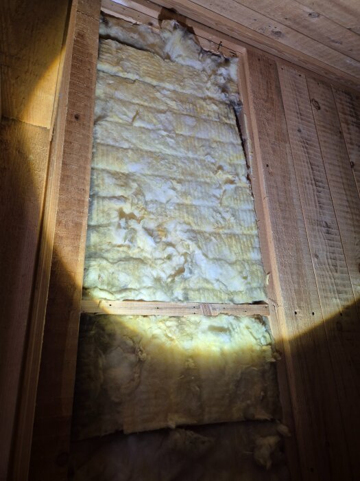 Inside view of a wall with insulation exposed, wooden beams, and gaps, illuminated by flashlight, possibly in an attic space of a wooden house.