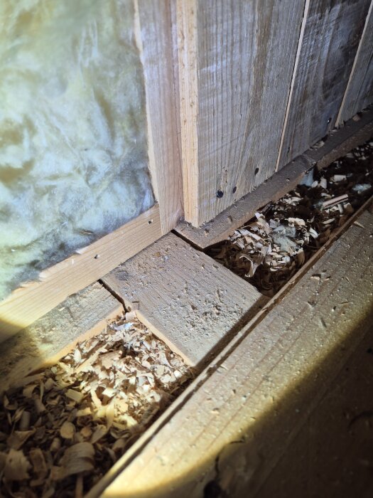 Interior view of attic slanted wall with insulation and wood shavings, showcasing construction materials and an uncovered section exposing structure details.