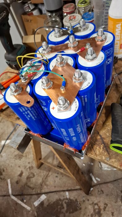 Blå battericeller med kopplingsplattor monterade i serie på en arbetsbänk, omgivna av verktyg och glasburkar.