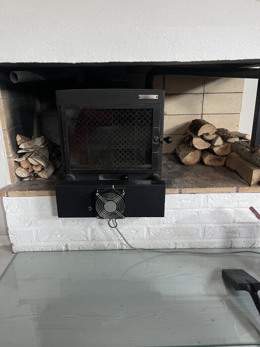 Gammal insatskamin i murad spis med glaslucka, ventilationsfläkt och vedträn bredvid.