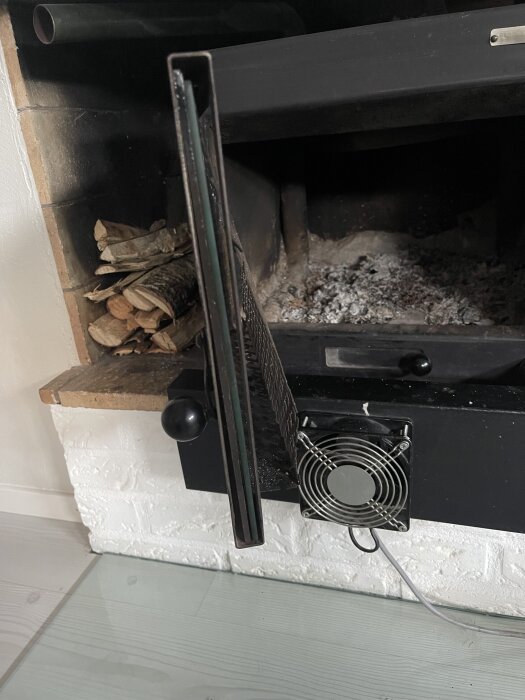 Öppen insatskamin med delvis öppen lucka, visar platsen där glaset tas ut och en synlig fläkt under. Ved syns bredvid kaminen.