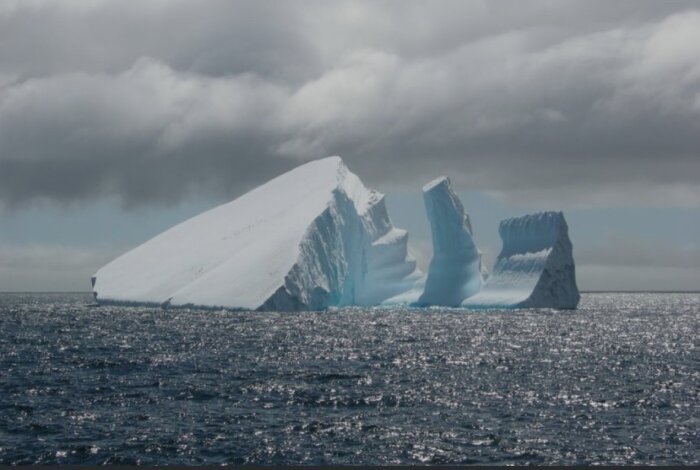 Stort isberg i havet under molnig himmel, med två mindre isberg synliga i bakgrunden.