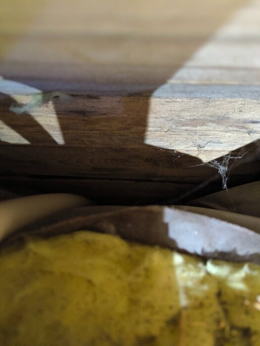 Isolering av glasfiber under trägolv med synligt damm och några rör. Skuggor och spindelväv är också synliga på bilden.