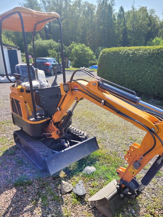 En orange minigrävare på en grusuppfart, omgiven av träd och buskar, under en solig dag.