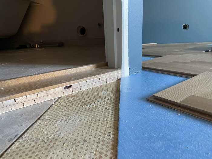 Höjd skillnad mellan badrumströskel och parkettgolv med synlig blå isolering och träbräda som sticker ut, lösning söks för att undvika gap.