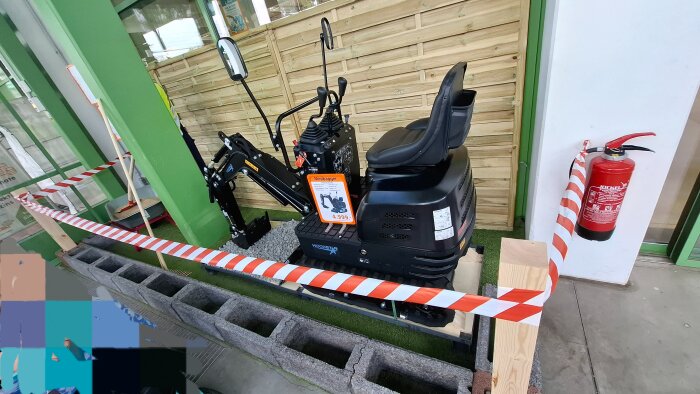 Minigrävare utställd i entré till tyskt byggvaruhus, omgiven av varningstejp och försedd med prisskylt.