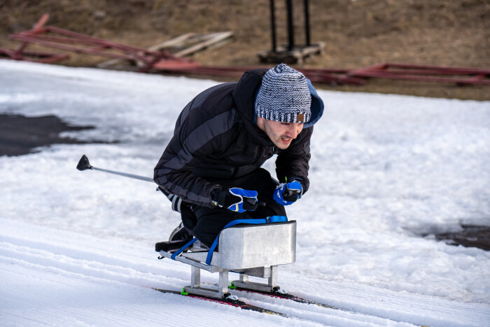 En person åker ett sitski på en snötäckt bana, klädd i vinterjacka och mössa, med stavar i händerna.