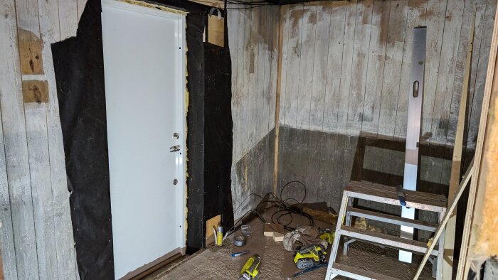 Under renovering av en gammal hästbox visas en nyinstallerad vit dörr, verktyg på golvet och en stege lutad mot väggen.