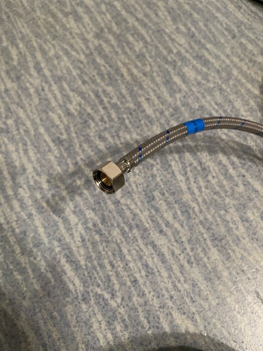 Flexibel metallslang med gängad anslutning, kanske en vattenanslutning till en blandare i badrum, visas på mönstrat underlag.