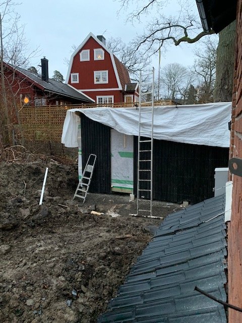 Ofärdigt byggprojekt med tak och väggar, utan dörrar. Två stegar står lutade mot den svarta strukturen täckt med presenning, omgivande träd och hus.