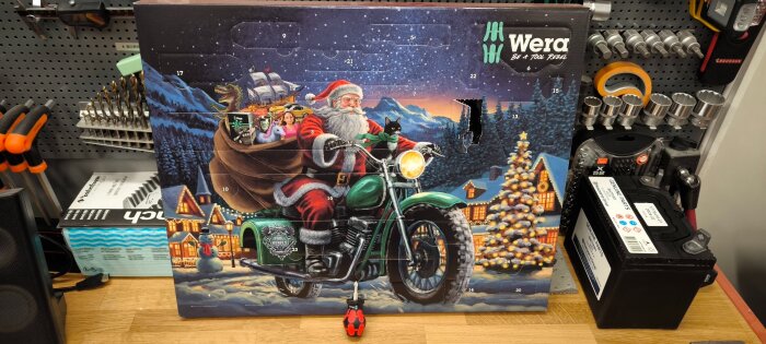 Julkalender med jultomten på en motorcykel i ett snölandskap. Kalendern har uppslagbara luckor och är omgiven av verktyg på en hylla.
