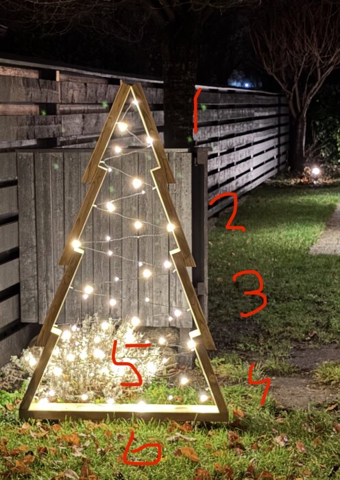 Trästruktur formad som en julgran med en ljusslinga, placerad på en gräsmatta vid ett trästaket.