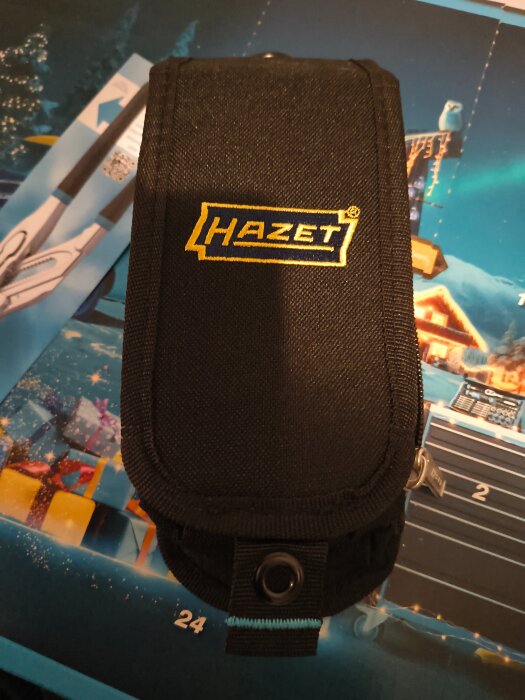 Svart väska med Hazet-logotypen i gult, placerad på en färgglad bakgrund med paketmotiv.