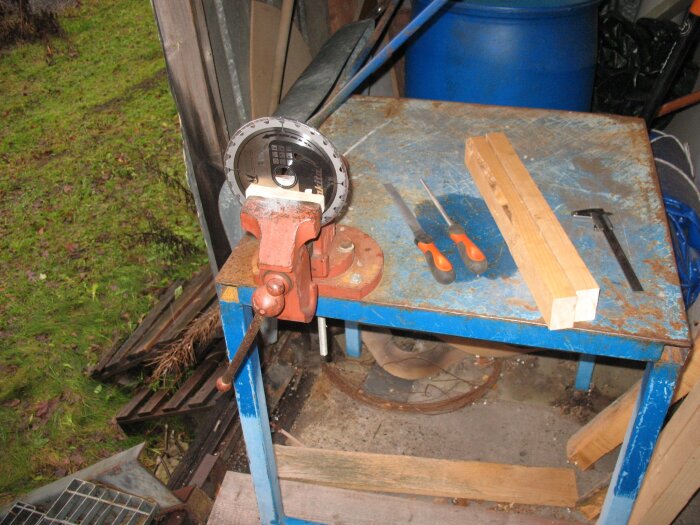 Handcirkelsåg i skruvstäd på verkstadsbord med verktyg som fil och skjutmått, omgivet av trä och skogsmark i bakgrunden.