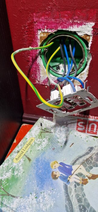 Väggkontakt med kablar där jordkabeln inte är ansluten, synlig röd vägg och bok med omslag av Astrid Lindgren.