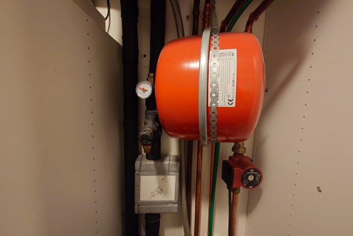 Rött expansionskärl ovanför en pump med manometer och rörsystem, installerat i ett skåp.