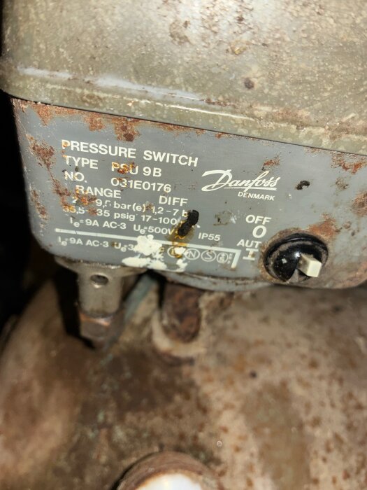Gammal tryckströmbrytare av typen Danfoss PSU 9B med rost och märkningar, monterad på kompressor, nära en avluftningsventil.