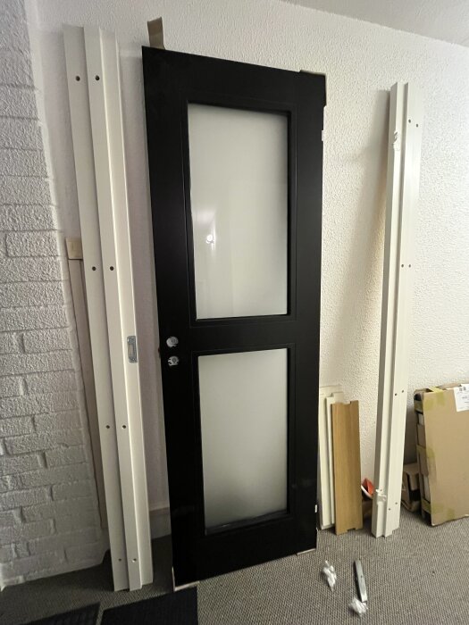 Svart dörr med karm och glaspartier mot vit vägg, omgiven av diverse byggmaterial.
