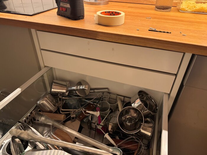 Öppen kökslåda i ett Ikea Metod-skåp, fylld med köksredskap och skålar, med en borrmaskin och skruvar på bänkskivan ovanför.