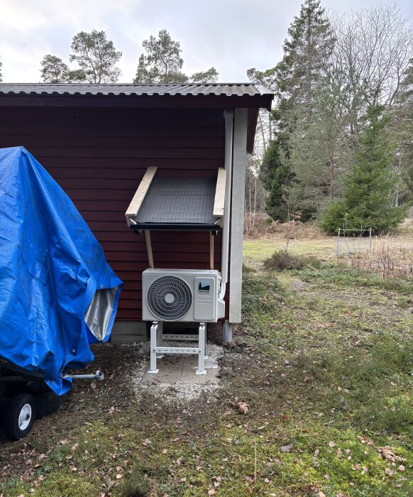 Litet tak av brädor och takpapp över en värmepump vid en röd byggnad, intill ett förråd täckt med blå presenning.