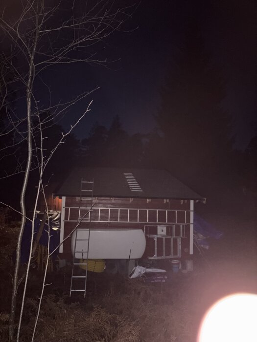 Byggnad under renovering i mörker, med stege och byggmaterial intill. En stor vit tank syns under byggnaden och en ensam gren syns till vänster.
