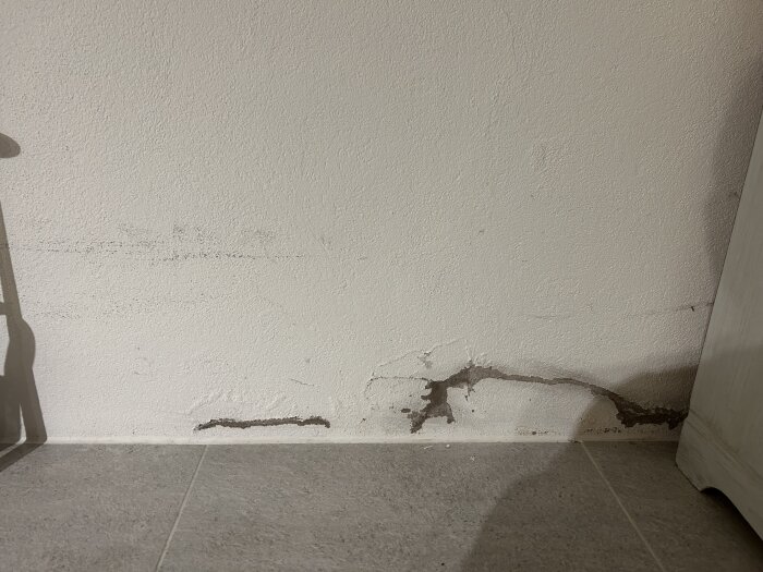 Fuktskada på en vit putsad innervägg i siporex, nära golvet, med mörka fläckar och sprickor synliga.