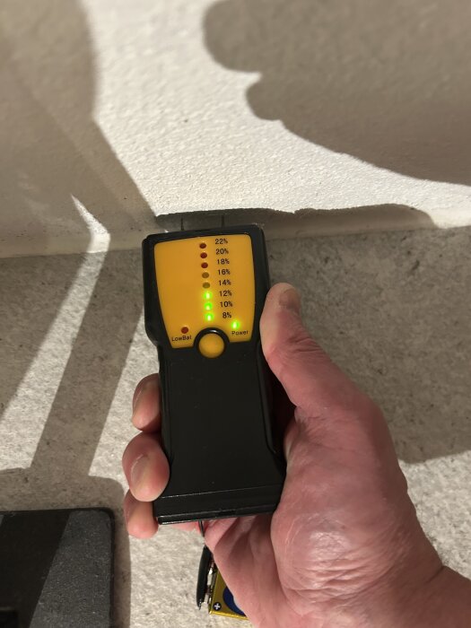 En hand håller en fuktmätare mot en murad innervägg, display visar 8% fuktnivå, lysande gröna lampor markerar mätningen.