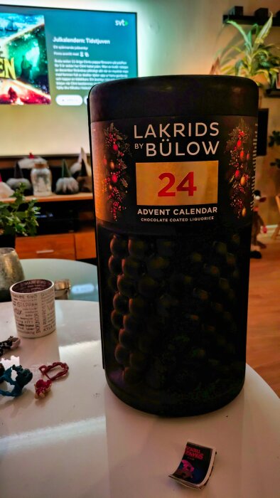 En stor burk med adventskalender "Lakrids by Bülow 24" står på ett bord, med en tv i bakgrunden som visar julkalendern på SVT.