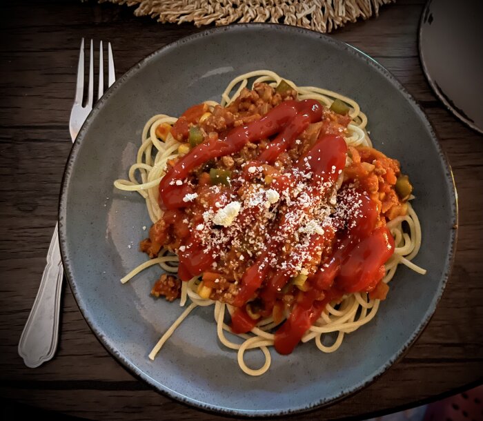 Spaghetti med köttfärssås, taco-inspirerad, toppad med ketchup, majs, paprika och riven ost. Serverad på tallrik med gaffel.