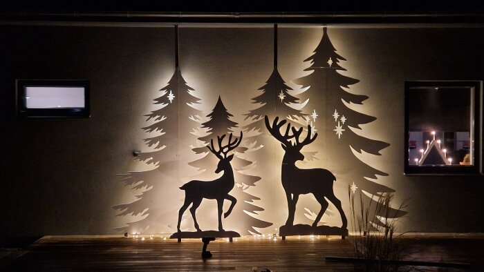 Silhuett av renar och granar som vinterdekoration, upplyst med ljusslingor mot en vägg.